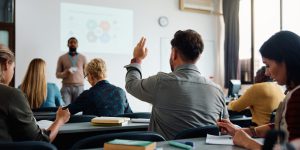 Rear view of man raising arm to ask a question during a presentation in lecture hall.
